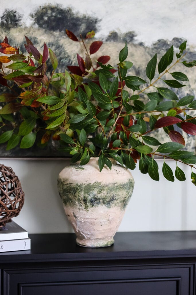 rustic looking vase filled with green branches on a mantle.
