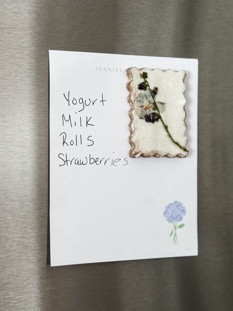 a short grocery list on fridge with a pressed flower magnet holding it in place
