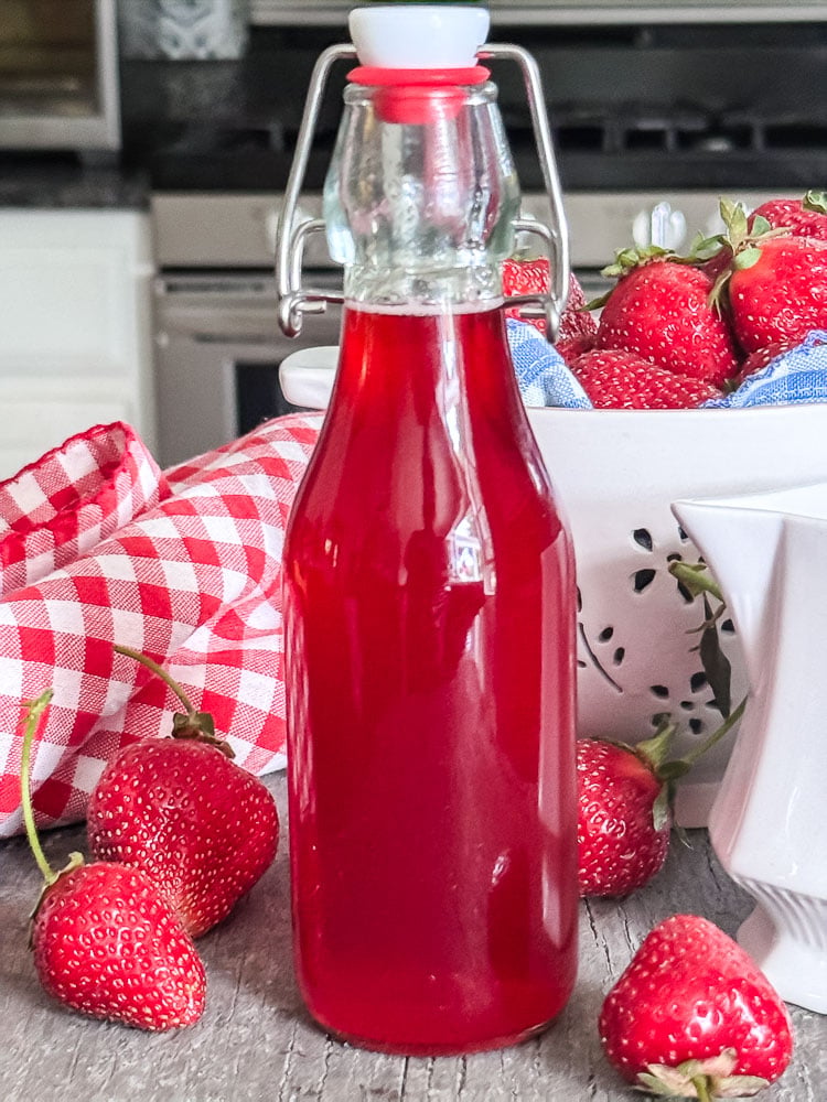 a glass blottle of strawberry syrup surrounded by fresh strawberries and a kitchen in the background