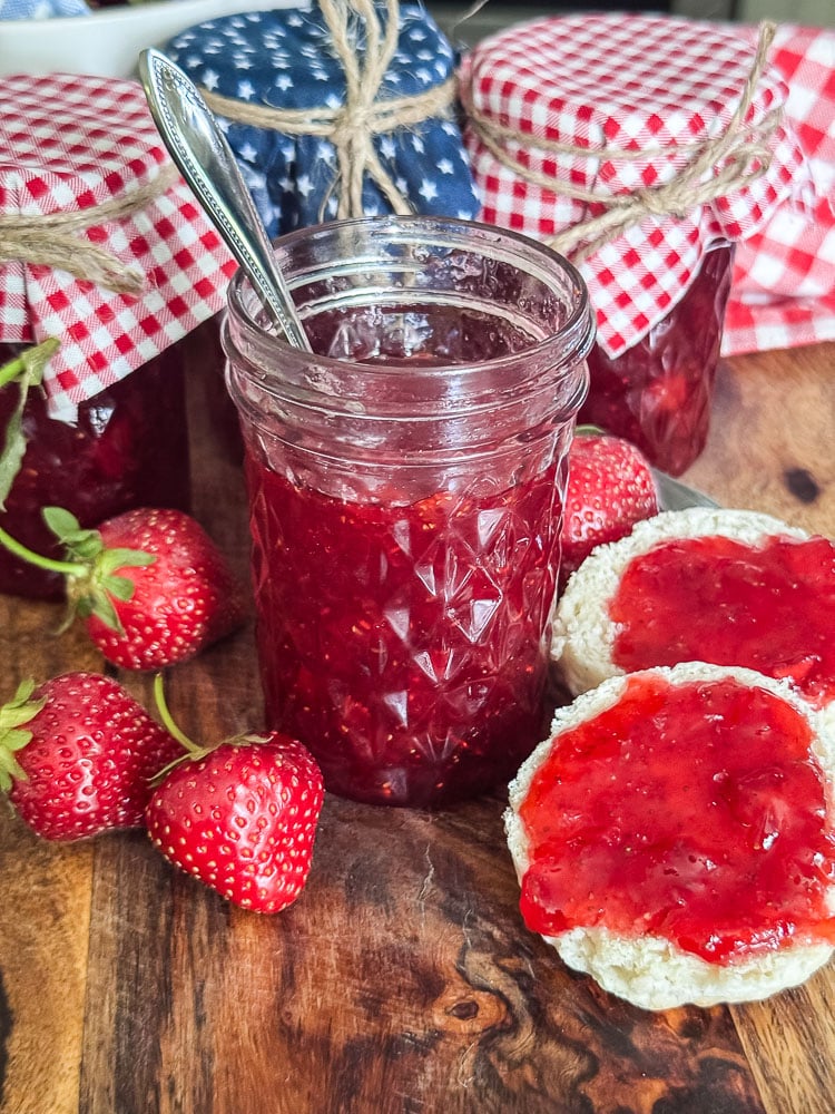 an opened jar of strawberry jam and a b iscuit with jam with more jars in background covered with fabric on the top