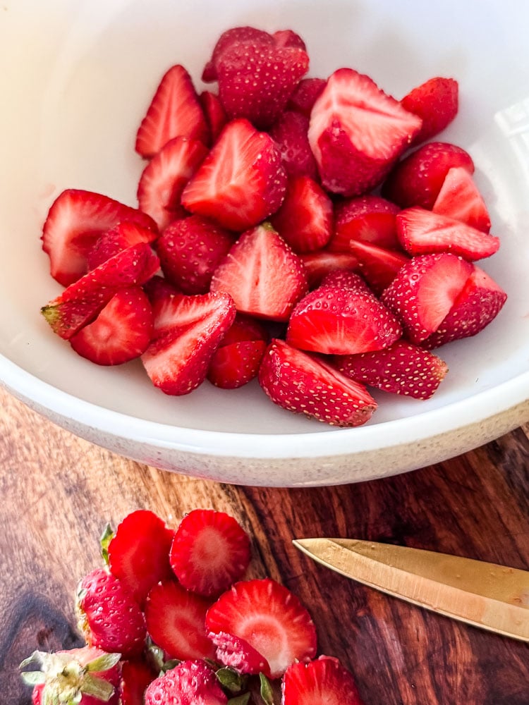 a bowl of halved strawberries on a cutting board with a paring knife and whole berries